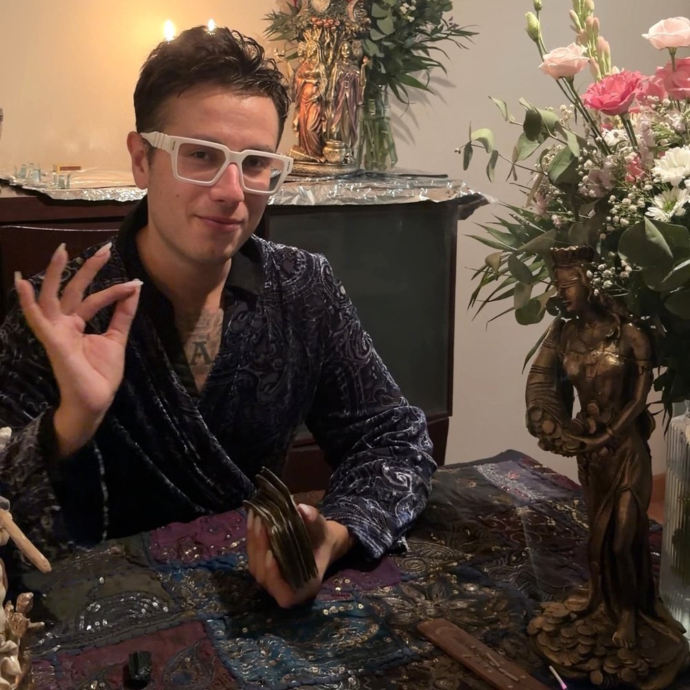 Alejandro, de Tarot de Encarna, con una baraja de cartas en la mano ante una mesa con flores y figuras ceremoniales